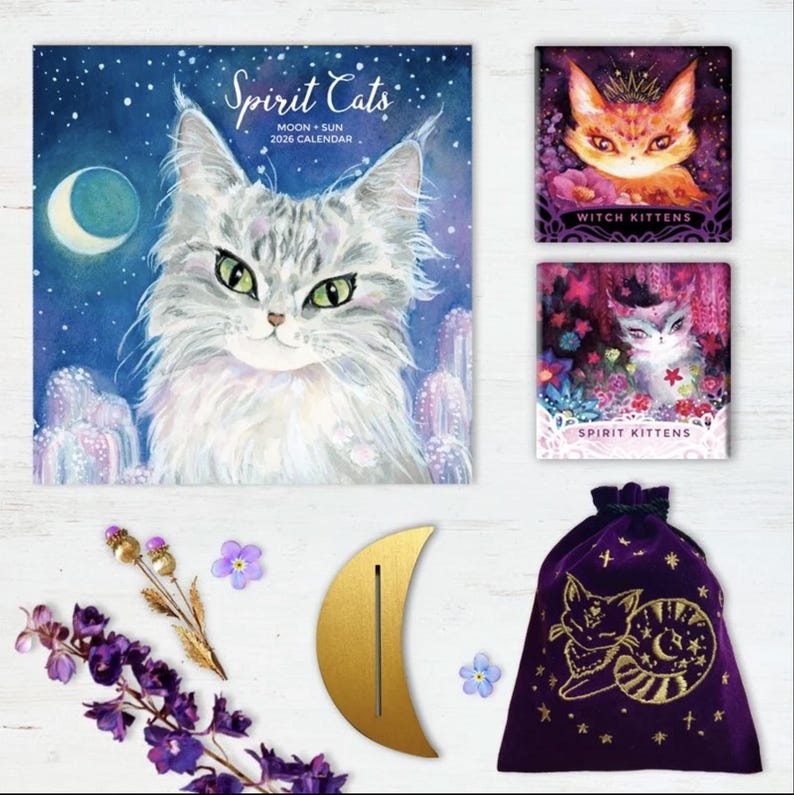 Może przedstawiać: Kolekcja przedmiot&oacute;w z motywem kota, w tym kalendarz "Spirit Cats" 2026, dwa wydruki "Witch Kittens" i "Spirit Kittens", złoty stojak w kształcie p&oacute;łksiężyca, fioletowy aksamitny woreczek i akcenty kwiatowe.