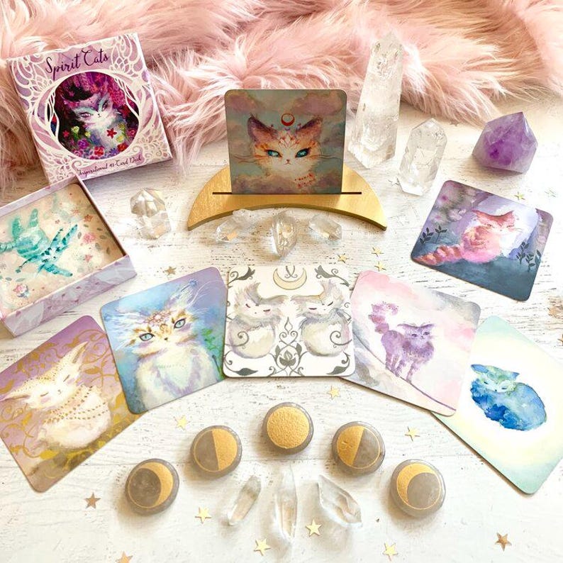 Pode incluir: Um conjunto de cartas de or&aacute;culo Spirit Cats com ilustra&ccedil;&otilde;es de gatos em v&aacute;rias poses. As cartas est&atilde;o dispostas sobre uma superf&iacute;cie branca com cristais e pedras. As cartas fazem parte de um conjunto maior de cartas de or&aacute;culo.