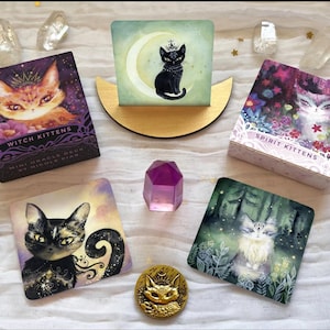 Może przedstawiać: Asortyment ilustrowanych kart tarota i przedmiot&oacute;w dekoracyjnych. Karty przedstawiają ilustracje kot&oacute;w z tytułami takimi jak "Witch Kittens" i "Spirit Kittens". Obecna jest r&oacute;wnież złota moneta z kocim obliczem i fioletowy kryształ, a także przezroczyste kryształy kwarcu.