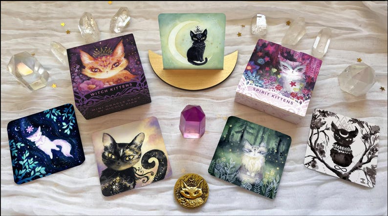 Może przedstawiać: Asortyment ilustrowanych kart tarota i przedmiot&oacute;w dekoracyjnych. Karty przedstawiają ilustracje kot&oacute;w z tytułami takimi jak "Witch Kittens" i "Spirit Kittens". Obecna jest r&oacute;wnież złota moneta z kocim obliczem i fioletowy kryształ, a także przezroczyste kryształy kwarcu.