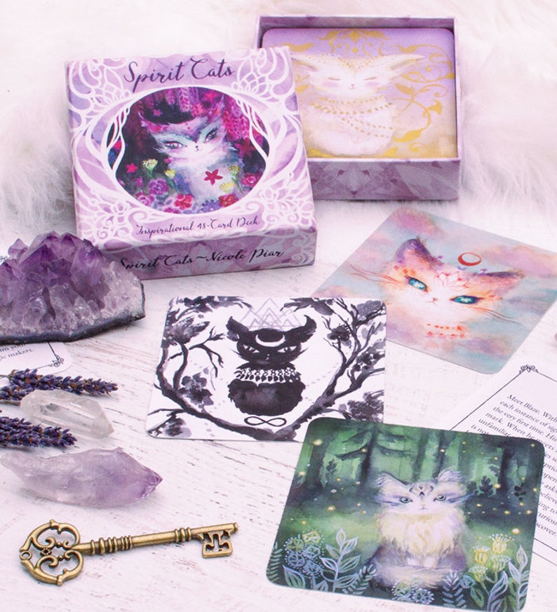 Pode incluir: Uma caixa roxa com o texto "Spirit Cats" e "Inspirational 48-Card Deck" na frente. A caixa cont&eacute;m um baralho de cartas com ilustra&ccedil;&otilde;es de gatos. As cartas est&atilde;o sobre uma superf&iacute;cie de madeira branca com cristais e uma chave.