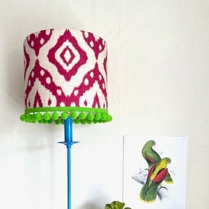 May include: A blue table lamp with a white and pink patterned lampshade with a green pom-pom trim. The lamp is on a wooden table with a stack of books and a small potted plant.