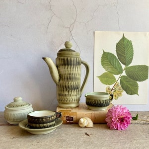 May include: A ceramic coffee set with a cream and black striped design. The set includes a coffee pot, creamer, sugar bowl, cup, and saucer. A botanical print and a pink flower are in the background.