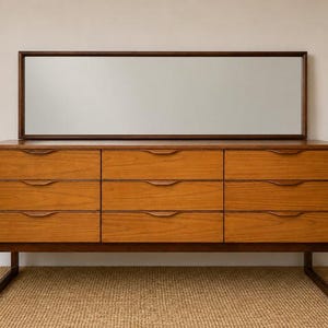 May include: A rectangular wooden chest of drawers featuring nine drawers arranged in three vertical columns. Each drawer has a carved horizontal handle. A large, rectangular mirror with a dark brown wooden frame is positioned directly above the unit.