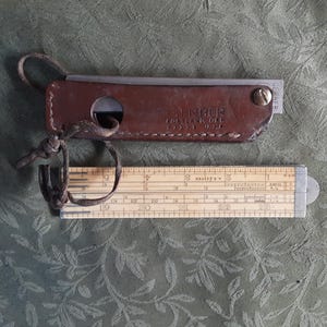Peut inclure: Une r&egrave;gle &agrave; calcul vintage et son &eacute;tui en cuir. L'&eacute;tui est marron et porte le mot "TIMBER" en relief. La r&egrave;gle &agrave; calcul est en bois et en m&eacute;tal, avec des mesures en centim&egrave;tres.