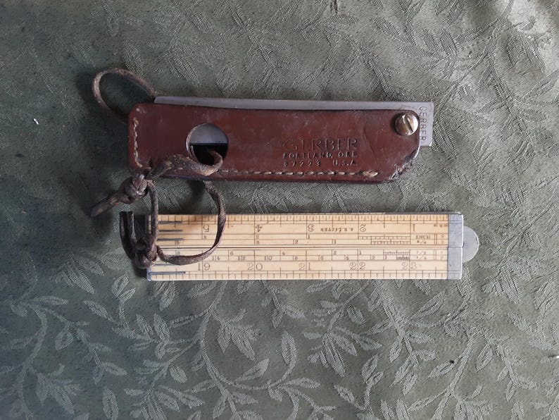 Peut inclure: Une r&egrave;gle &agrave; calcul vintage et son &eacute;tui en cuir. L'&eacute;tui est marron et porte le mot "TIMBER" en relief. La r&egrave;gle &agrave; calcul est en bois et en m&eacute;tal, avec des mesures en centim&egrave;tres.