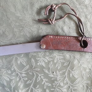 Peut inclure: Un outil vintage en cuir et m&eacute;tal. Il pr&eacute;sente une poign&eacute;e en cuir marron et une lame argent&eacute;e. La poign&eacute;e a un design cousu et un petit trou. Une lani&egrave;re en cuir est attach&eacute;e &agrave; la poign&eacute;e.