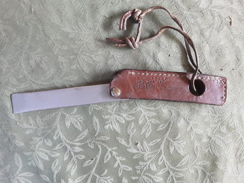 Peut inclure: Un outil vintage en cuir et m&eacute;tal. Il pr&eacute;sente une poign&eacute;e en cuir marron et une lame argent&eacute;e. La poign&eacute;e a un design cousu et un petit trou. Une lani&egrave;re en cuir est attach&eacute;e &agrave; la poign&eacute;e.
