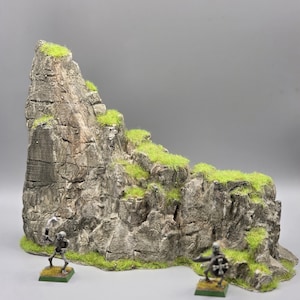 Può includere: Una dettagliata formazione rocciosa in miniatura con ciuffi d'erba verde, adatta per giochi da tavolo. Due piccole figure di scheletri sono alla base. La roccia è grigia con dettagli strutturati, creando un paesaggio realistico.