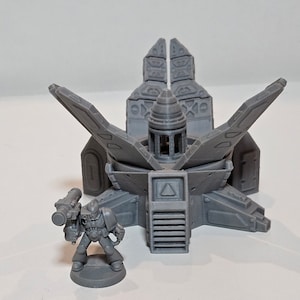 May include: A gray, 3D-printed miniature model of a futuristic structure with a central spire and wing-like extensions. A small, gray figurine of a soldier with a weapon stands nearby. The model has intricate details and a geometric design.