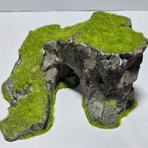 Op de afbeelding: Een miniatuur rotsformatie met een natuurlijke boog, bedekt met levendig groen kunstgras. De grijze steenstructuur heeft een textuur, waardoor een realistisch landschapselement ontstaat voor modeldisplays of terrariums.