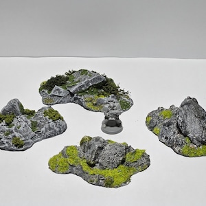 Könnte beinhalten: Vier graue Miniatur-Felsformationen mit grünem Moos und Vegetation. Eine kleine graue Figur ist zwischen den Felsen positioniert. Ideal für Tabletop-Spiele oder Miniatur-Landschaften.