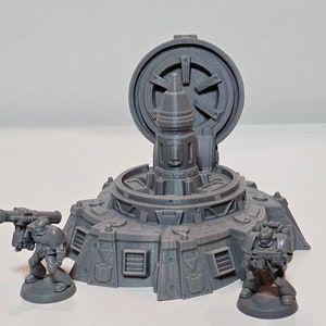 Könnte beinhalten: Graues, 3D-gedrucktes Miniaturmodell einer futuristischen Struktur mit kreisförmiger Oberseite und zentralem Turm. Zwei Miniaturfiguren stehen in der Nähe, eine hält eine Waffe. Das Modell hat detaillierte geometrische Designs.
