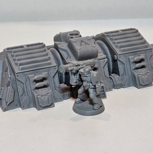 May include: A grey, 3D-printed miniature model of a futuristic structure with a soldier figure holding a weapon. The structure has a detailed, industrial design with rectangular and cylindrical components.