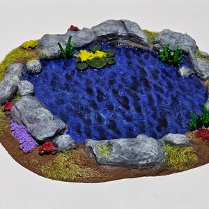 May include: A miniature pond diorama with a blue water surface, surrounded by grey rocks, green grass, and colourful plants. Yellow lily pads float on the water. The pond is a detailed, handcrafted piece.