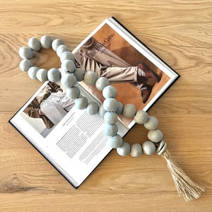 May include: A long strand of light gray wooden beads with a natural fiber tassel. The beads are strung together on a thick cord. The beads are round and have a smooth, matte finish. The tassel is about 6 inches long and is made of natural fibers. The beads are lying on top of a magazine with the text "The Polo Chino" visible.