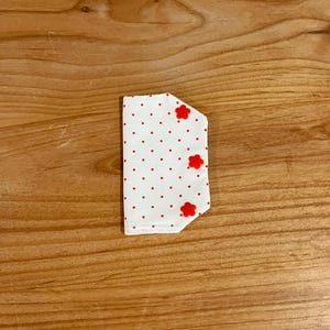 Peut inclure: Petit objet en tissu blanc à pois rouges et trois motifs rouges en forme de fleur. L'objet est rectangulaire avec un bord incurvé, posé sur une surface en bois.