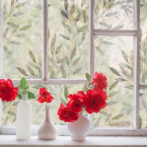 May include: A window with a leafy patterned film, featuring red roses in three white vases. The roses are arranged in a bouquet and the vases are on a white windowsill.