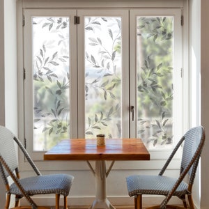 Puede incluir: Una pequeña mesa de madera con dos sillas se encuentra frente a una ventana con un film de privacidad con estampado floral. Las sillas son de mimbre con cojines azules.