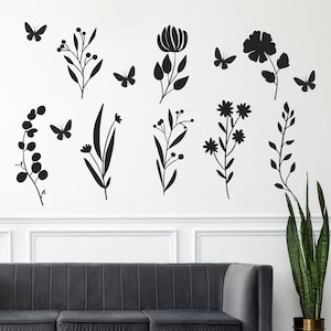 Puede incluir: Calcomanías de pared en silueta negra y blanca con varios diseños florales y de mariposas. Las calcomanías están dispuestas en una pared blanca con un sofá gris en primer plano.