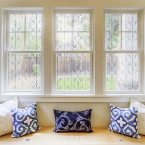 May include: Three windows featuring a semi-transparent frosted film with a repeating white leaf pattern. Below the windows, a wooden bench seat is adorned with two solid white square pillows, two blue and white patterned square pillows, and one rectangular dark blue pillow with a white floral design.
