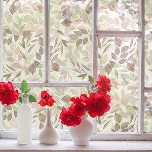 May include: Three white ceramic vases containing vibrant red roses are positioned on a window sill. The window glass features a semi-transparent decorative film with a repeating pattern of soft green and brown leaves across the entire pane.