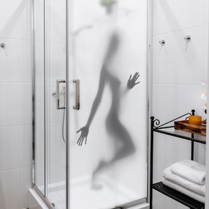 Könnte beinhalten: Eine Duschkabine mit einer Milchglastür, die die Silhouette einer Person zeigt. Das Badezimmer hat weiße Fliesen. Ein schwarzes Metallregal enthält Handtücher und ein Tablett mit einer Kerze.