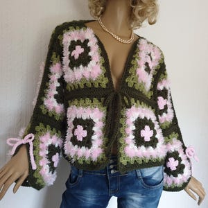 May include: A handmade, open-front crochet jacket in a granny square pattern. The jacket features olive green, white, and pink squares with fluffy trim. The sleeves have pink bows at the cuffs. The jacket is displayed on a mannequin.