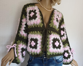 Puede incluir: Chaqueta de crochet hecha a mano, abierta por delante, con un patrón de cuadrados de la abuela. La chaqueta presenta cuadrados verde oliva, blancos y rosas con ribete esponjoso. Las mangas tienen lazos rosas en los puños. La chaqueta se muestra en un maniquí.