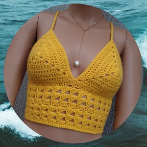 Puede incluir: Un top corto de crochet amarillo con un escote en V profundo y un delicado patrón de punto abierto. El top se lleva en un maniquí sobre un fondo de agua azul.