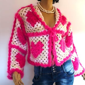 May include: A handmade, open-front crochet jacket in a granny square pattern. The jacket features a vibrant pink heart design on a cream and beige background, with fluffy pink trim. The sleeves are long, and the jacket is worn over blue jeans.
