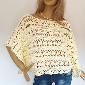 May include: A cream-coloured crochet top featuring an openwork pattern with repeating shell-like motifs and horizontal bands. The garment has a wide, loose-fitting silhouette with short, flared sleeves and a rounded neckline, crafted from yarn.