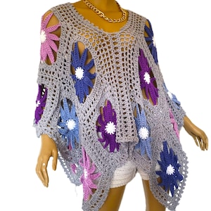 May include: A gray crochet poncho with large, colorful flower cutouts in shades of purple, blue, and pink. The poncho has a V-neck and a loose, flowing design, perfect for layering. The flowers have white centers.