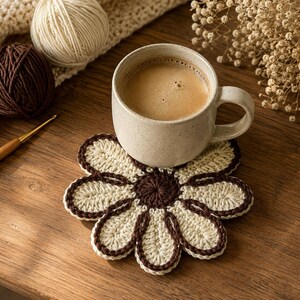 May include: A round, flower-shaped crochet coaster featuring a dark brown center and eight cream-colored petals outlined in dark brown yarn. A speckled beige ceramic mug filled with coffee sits directly on top of the coaster.