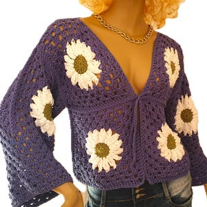 May include: A periwinkle blue crochet cardigan with large white and green daisy appliqués. The open-front sweater has long sleeves and a V-neckline. The daisy details are placed on the front and sleeves.