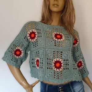 May include: A light teal crochet top with a floral pattern. The top features a square design with red and orange floral accents. The sleeves are three-quarter length, and the neckline and hem have a scalloped edge.
