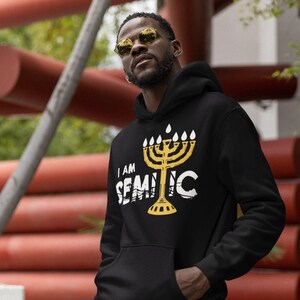 May include: Black hoodie with a gold menorah graphic and the text "I AM SEMITIC". The hoodie has a front pocket and a fringed hem. The design is a statement of identity and cultural pride.
