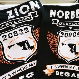 May include: Two black t-shirts with white text and graphics. The left shirt says "MT Zion Maryland 20832" with a graphic of the state of Maryland and wings. The right shirt says "Norbeck Maryland 20906" with a graphic of the state of Maryland and wings. Both shirts have a banner that says "It's where my story began."