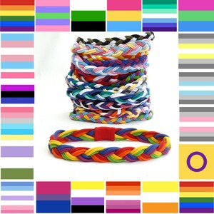 May include: A collection of braided hairbands in a variety of colours, including rainbow, pink, blue, and green. A single rainbow hairband with a red clasp is at the bottom. Pride flags are arranged around the hairbands.
