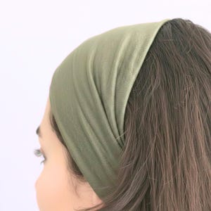 May include: An olive green headband made from a soft, stretchy material. The wide band is worn across the forehead, covering the hairline. The headband is a solid colour and appears to be knitted. The hair is brown and long.