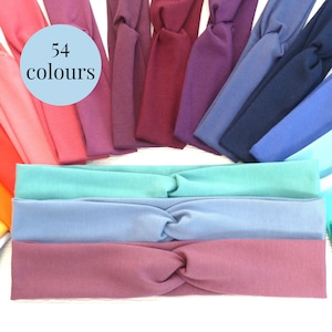 K&ouml;nnte beinhalten: Eine Auswahl an Stirnb&auml;ndern in verschiedenen Farben, darunter Rosa, Lila, Blau und Gr&uuml;n. Das Bild zeigt einen hellblauen Kreis mit dem Text "54 Farben". Die Stirnb&auml;nder sind aus weichem Stoff gefertigt und haben ein gedrehtes Design.