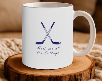 May include: A white ceramic mug featuring a dark blue graphic of two crossed hockey sticks. Below the sticks, the text "Meet me at the Cottage" is printed in a dark blue script font. The mug has a standard handle and a smooth, cylindrical body.