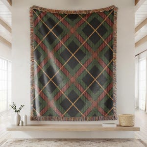 May include: A woven throw blanket featuring a large-scale diamond plaid pattern in dark charcoal, forest green, and muted red, accented by thin yellow diagonal lines. The rectangular textile has a decorative fringe along all four edges.