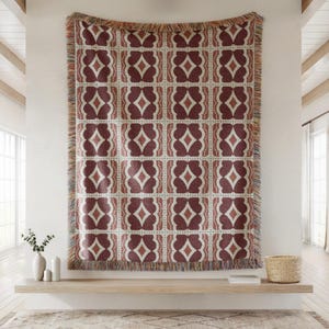 May include: A woven throw blanket featuring a repeating geometric pattern in deep maroon and cream. The design consists of symmetrical diamond and petal shapes arranged in a grid, finished with a multicolored fringed edge on all four sides.