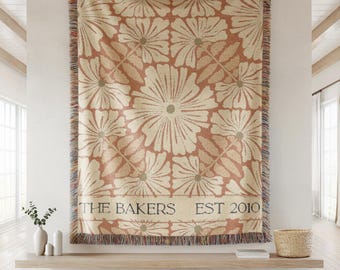 Pode incluir: Uma manta tecida com um grande padrão floral em tons de creme e pêssego. A manta tem uma borda com franjas e o texto "THE BAKERS EST 2010" é exibido na parte inferior. Pendurada em uma parede branca.