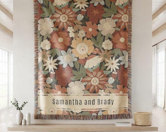 Pode incluir: Uma manta tecida com fundo escuro e um padrão floral multicolorido em tons de ferrugem, creme e verde sálvia. A secção inferior inclui uma faixa horizontal com o texto Samantha and Brady. As extremidades possuem franjas.