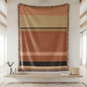 Op de afbeelding: Een geweven plaid met horizontale strepen in tinten terracotta, beige en crème, geaccentueerd door een dunne zwarte lijn. Het rechthoekige textiel heeft een getextureerd uiterlijk en is afgewerkt met decoratieve franjes langs alle vier de randen.