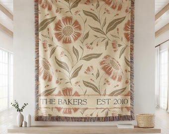 Pode incluir: Uma manta tecida com fundo creme e um padrão floral repetitivo em tons de terracota e verde sálvia. A secção inferior inclui uma borda horizontal com o texto THE BAKERS EST 2010. As extremidades apresentam franjas multicoloridas.