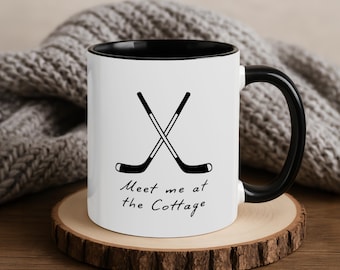 May include: White ceramic mug with a black interior and handle. The mug features a crossed hockey stick graphic and the text "Meet me at the Cottage" in a script font. The mug sits on a wooden coaster.