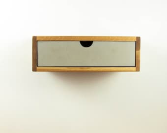 May include: A small, wooden wall-mounted shelf with a single drawer. The drawer is made of light grey concrete and has a black knob. The shelf is mounted on a white wall.